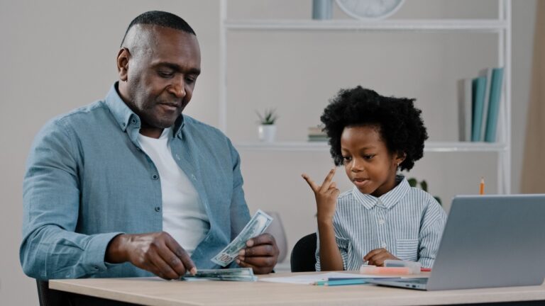 Cheerful adult father and little cute daughter sitting at home dad teaches kid girl to count shows dollars