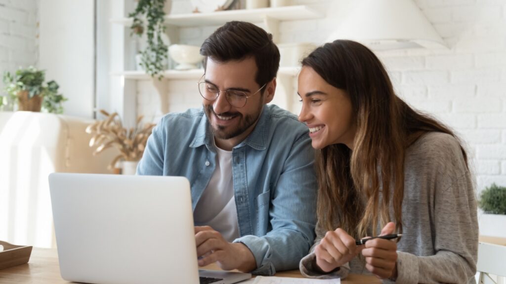 Happy young couple husband and wife using laptop computer looking at screen pay bills online in app calculate mortgage investment payment on website planning budget discuss finances sit at home table