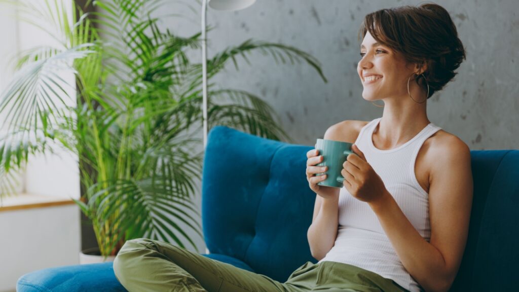 Side view young woman wears white tank shirt drink tea coffee sit on blue sofa couch stay at home hotel flat rest relax spend free spare time in living room indoors grey wall. People lounge concept
