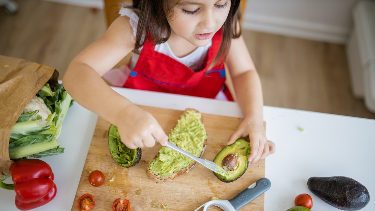 Adorable little girl at white table spreading avocado on slice of bread.