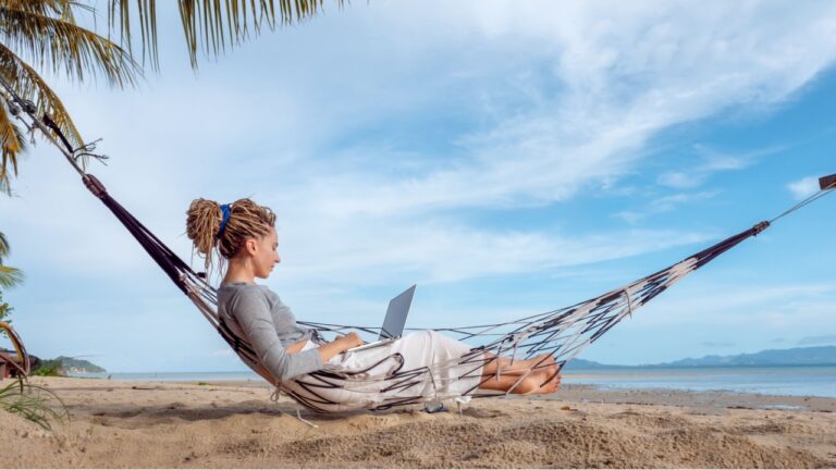 Young woman working on laptop lying in hammock at sand beach of tropical island.